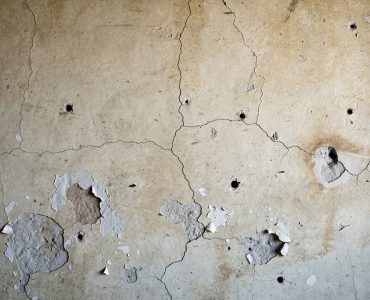 Mur intérieur ancien présentant de petites fissures, des trous rebouchés et des zones de peinture écaillée, éclairé par une lumière naturelle latérale.