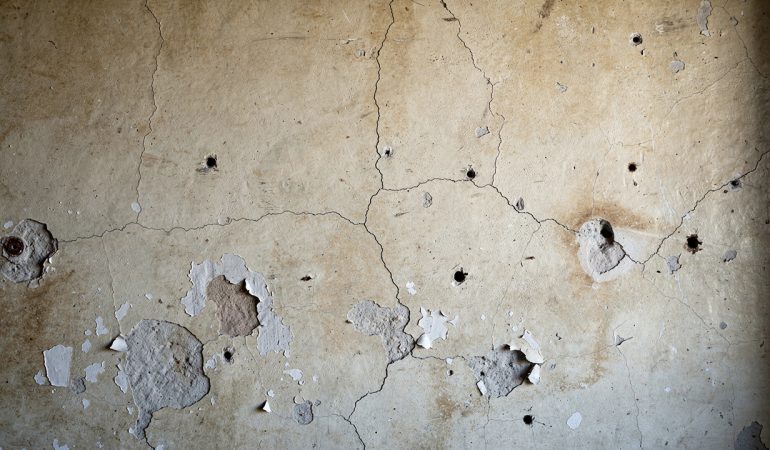 Mur intérieur ancien présentant de petites fissures, des trous rebouchés et des zones de peinture écaillée, éclairé par une lumière naturelle latérale.
