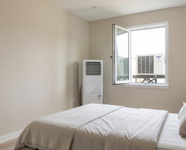 Chambre lumineuse avec un climatiseur mobile split près d’une fenêtre entrouverte et calfeutrée, dont l’unité extérieure repose sur un support sécurisé.