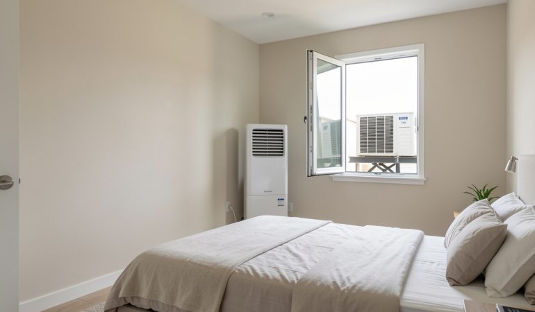 Chambre lumineuse avec un climatiseur mobile split près d’une fenêtre entrouverte et calfeutrée, dont l’unité extérieure repose sur un support sécurisé.